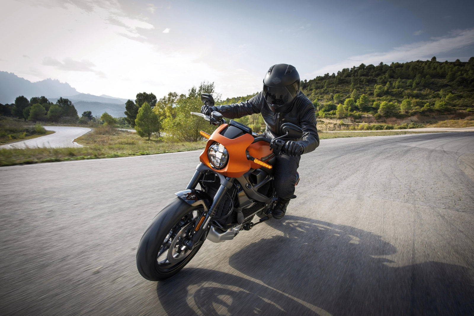 Stunt rider, obscured by a black helmet and leather jacket, dominates the center of the frame as he traverses a winding road